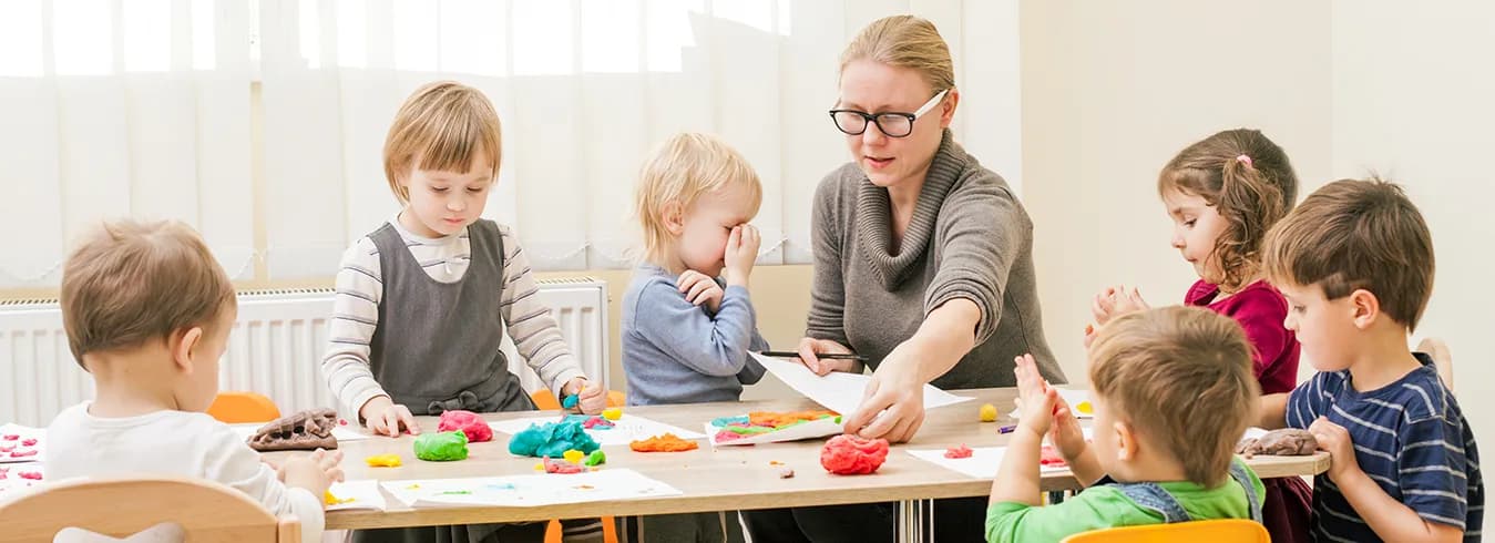 A teacher and a group of preschoolers seated around a classroom table, modelling shapes out of brightly coloured play dough.