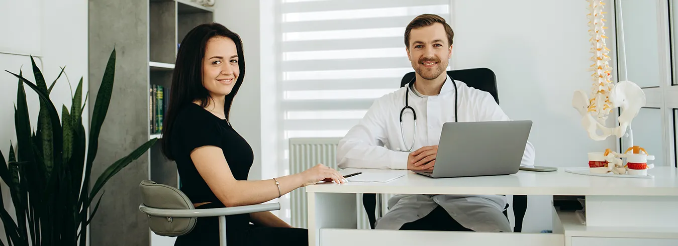 A doctor in a white coat and stethoscope working at a clinic desk beside a seated patient, with an anatomical spine model behind them.