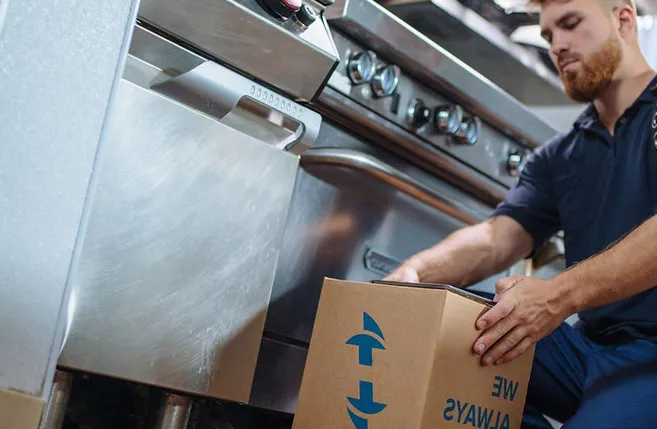 A service technician in uniform kneeling beside a stainless commercial range, unpacking a parts box