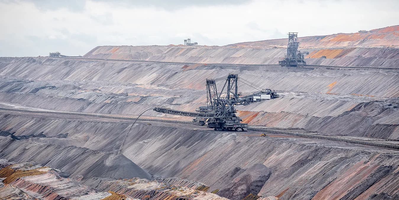 An open-pit mine under an overcast sky: two bucket-wheel excavators working terraced benches of grey and rust-coloured overburden, with a conveyor line running across the pit floor.