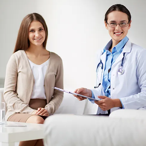 A doctor in a white coat holding a clipboard, seated beside a patient in a bright consultation room.