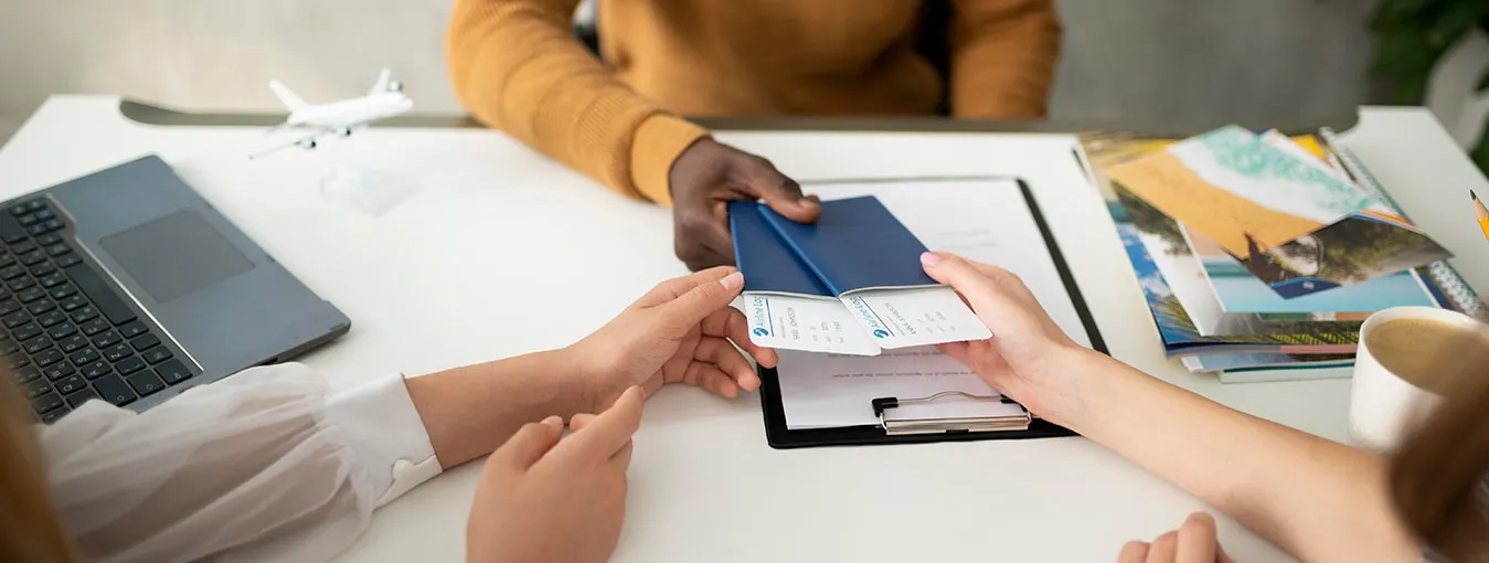 An immigration consultant handing a passport and travel documents across a desk to a client, beside a laptop and brochures.