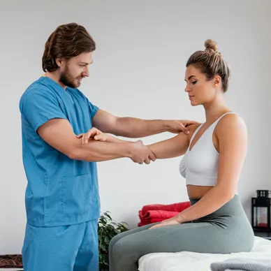 A physiotherapist in blue scrubs stretching a seated patient’s arm during a treatment session.