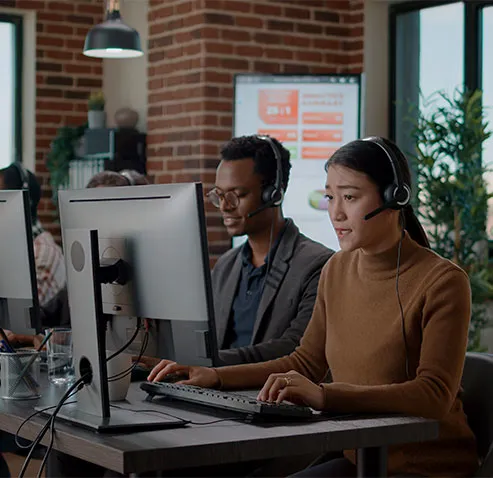 Two support agents wearing headsets working at desktop monitors in a brick-walled office.